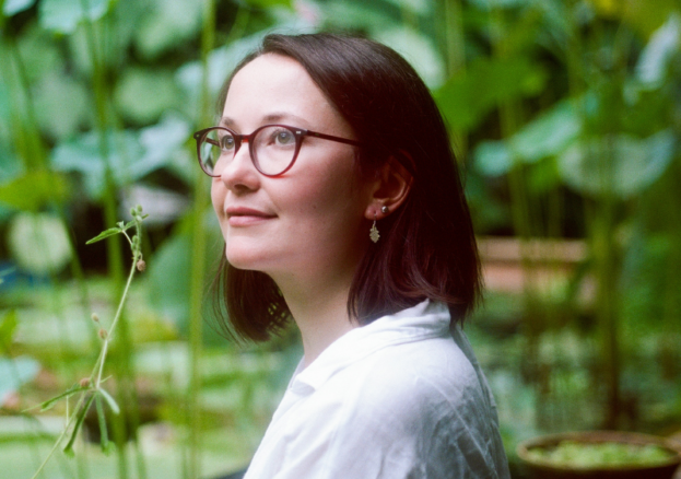 An image of author Marianne Brooker stood in front of green fauna, wearing glasses, smiling and looking up away from camera.