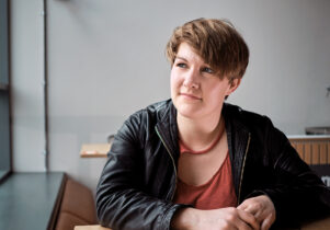 Portrait of Caroine in a black leather jacket and rust coloured tshirt. She has white skin and short brown hair sweeping acrcoss her face down to the right