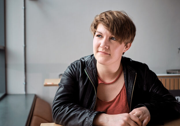 Portrait of Caroine in a black leather jacket and rust coloured tshirt. She has white skin and short brown hair sweeping acrcoss her face down to the right