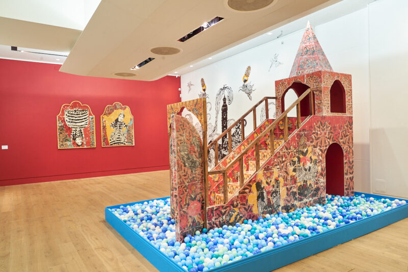 A gallery installation featuring a structure with steps up to a tower with a pointed roof, surrounded by a children's ball pit with balls of varying shades of blue. Two framed works in vibrant colours hang on a red wall behind it
