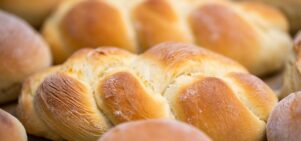 A close-up image of freshly baked challah bread. The bread is golden-brown, braided, and has a light, fluffy texture. Multiple loaves are visible in the background, all exhibiting a warm, inviting appearance with a slight dusting of flour.