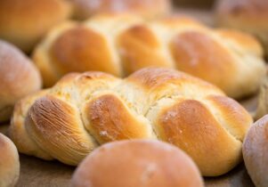 A close-up image of freshly baked challah bread. The bread is golden-brown, braided, and has a light, fluffy texture. Multiple loaves are visible in the background, all exhibiting a warm, inviting appearance with a slight dusting of flour.