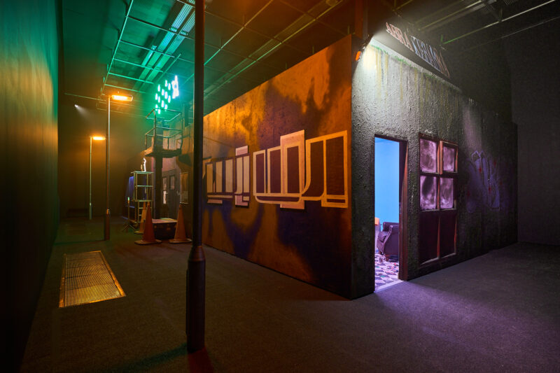 Image of a dark gallery with a building inside. Graffiti is on the walls and there is a street light in the foreground. Green neon signs hang above the building on the left side of the image.