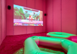 Image of a pink room with fluffy carpet and padded walls. A projection displays a scene from a videogame. In the foreground there are two large ringed inflatables that are lit with a green light from above. 