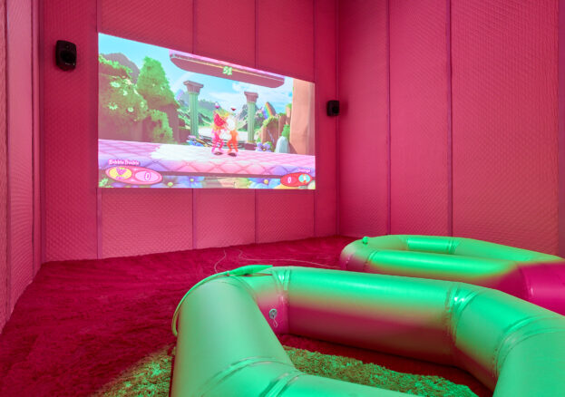 Image of a pink room with fluffy carpet and padded walls. A projection displays a scene from a videogame. In the foreground there are two large ringed inflatables that are lit with a green light from above. 