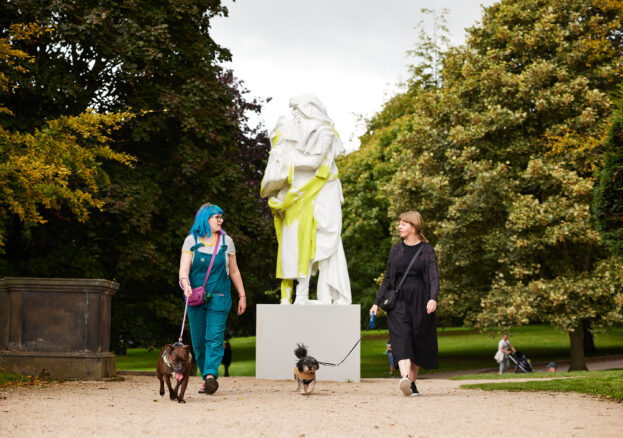 Two people walking their dogs in front of a sculpture