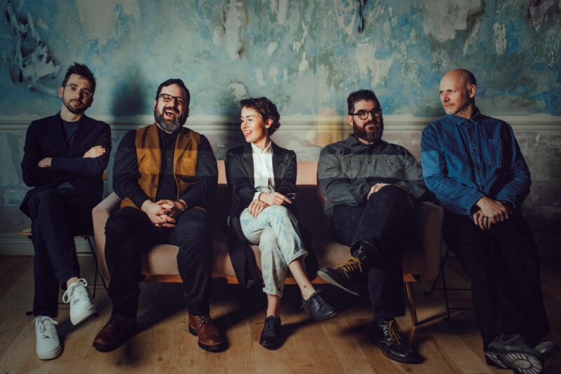 Five musicians sitting on a couch, four men and one woman in the middle