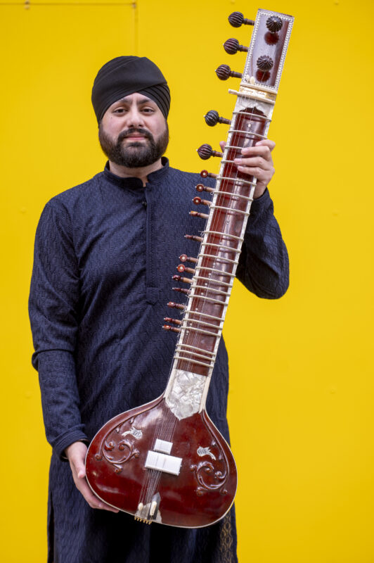 Male with beard holding sitar on with yellow background