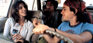 A young man, in the driver's seat of a car, passes a cigarette to the older woman next to him, while anther young man sits behind.