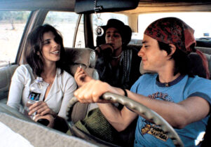 A young man, in the driver's seat of a car, passes a cigarette to the older woman next to him, while anther young man sits behind.