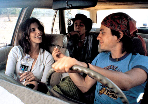 A young man, in the driver's seat of a car, passes a cigarette to the older woman next to him, while anther young man sits behind.