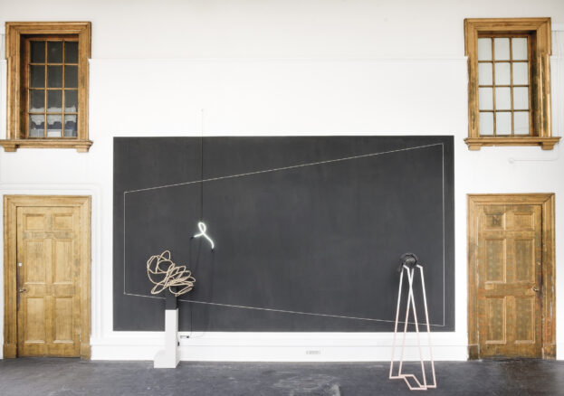 A minimalist art installation features geometric wire sculptures, a neon light in front of a black rectangle made of gesso, and two wooden doors with windows on either side in a white-walled room.