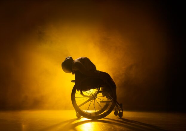 Performer in Wheelchair lit from behind. Lived Fiction at The Lowry