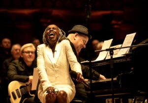 Female with white suit sitting on piano stool singing 