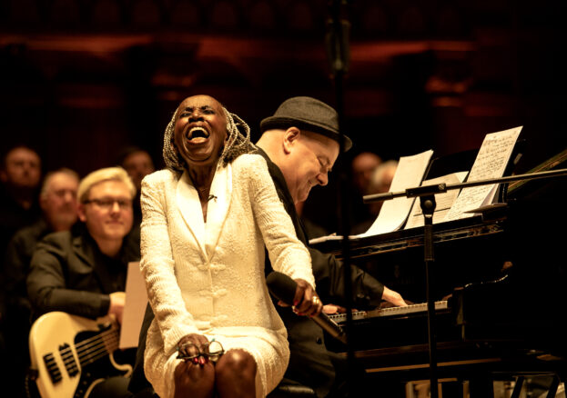 Female with white suit sitting on piano stool singing 