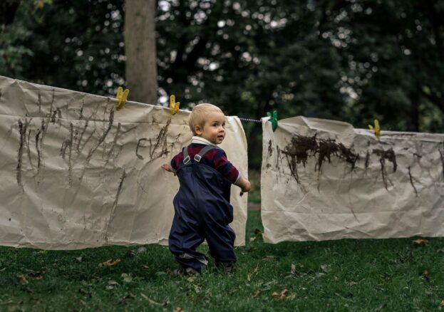 Toddler standing on grass with a paintbrush in hand turning to the right to look at the camera. He is wearing a navy blue rainsuit with a burgundy and navy blue long sleeved tiop underneath. He is standing in front of large cream coloured sheets that have been strung up on a washing line. The sheets have mud marks making on.