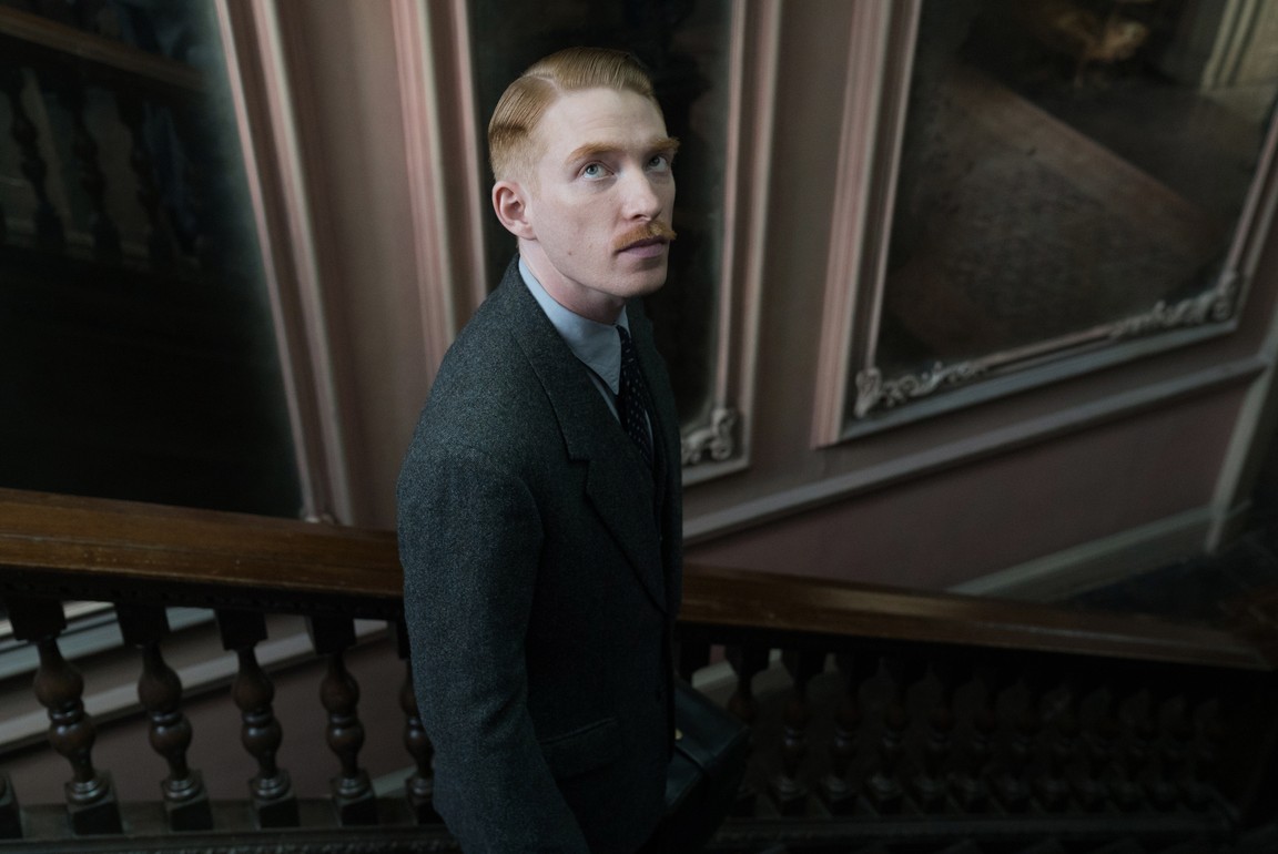 A moustached man in a large, old house, dressed smartly, stands on a staircase, looking up.