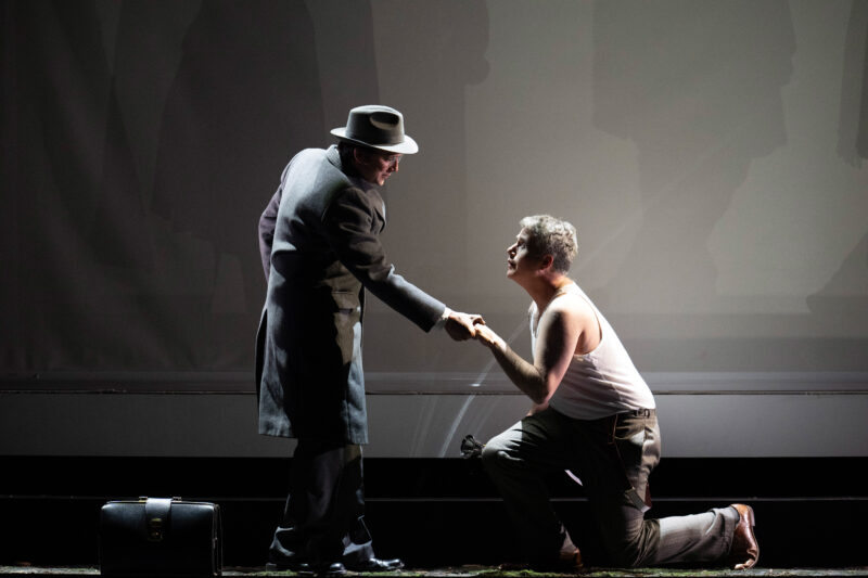 Blond Eckbert production image. Dark stage background with one man stood holding hand out to man on knees.