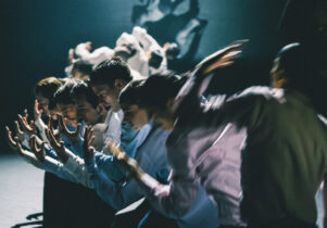 Figures in Extinction at Aviva Studios: A group of dancers, dressed in formal shirts, perform in unison on a dimly lit stage.