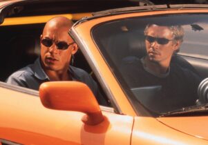 Vin deisel and Paul Walker wear sunglasses sitting in an orange sports car with the hood down