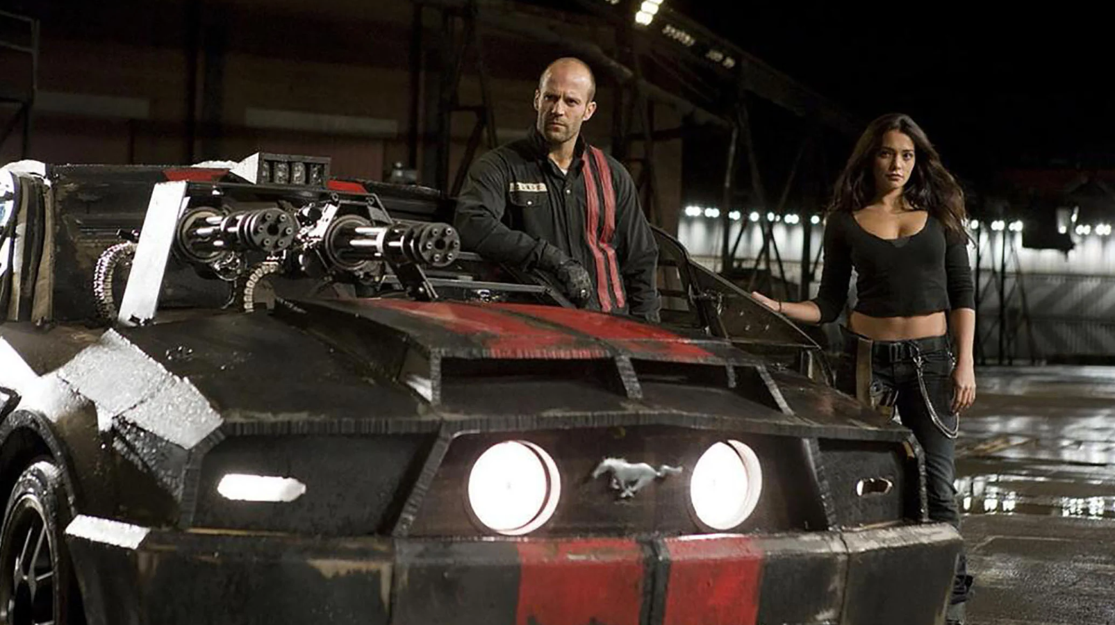 A man and a woman stand next to a red and black supercar with two large automatic weapons attached to the bonnet