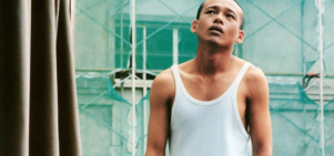 A man stands in the middle of shot infront of scaffolding wearing a white vest