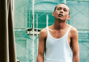 A man stands in the middle of shot infront of scaffolding wearing a white vest