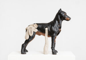 A sculpture of a dark brown dog looks to the right, hanging out of its middle and the back are what appears to be its insides (in cream) spilling out.