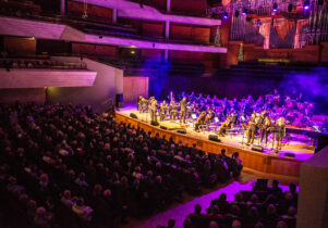 A concert hall stage with orchestra and audience, lit up in purple and blue lights