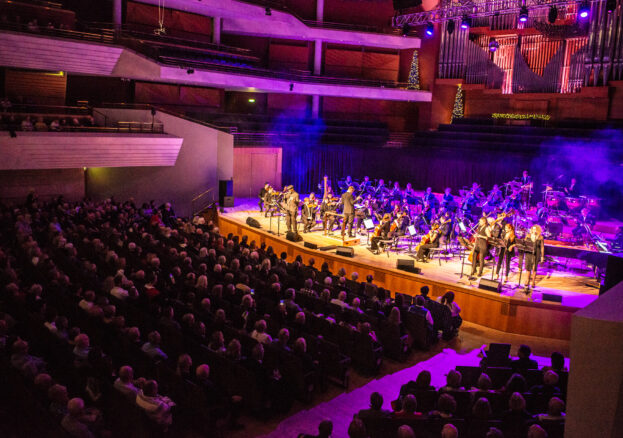 A concert hall stage with orchestra and audience, lit up in purple and blue lights