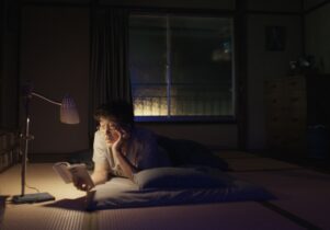 A man lies in bed reading a book by lamplight.