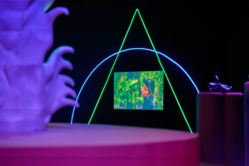 Blurred, purple lit sculpture in the foreground. At the centre of the image, there is a floating screen displaying a woman with dark hair amongst a forrest, embracing a tree. The screen is surrounded by neon lights, one in the shape of a green triangle, the other a blue semi-circle.