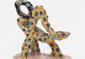 A brightly coloured abstract ceramic sculpture. Almost figure-like - a series of thin, round elements with yellow background and brown and blue spots, sit on a pink base, with a dark brown head-shaped piece on top.