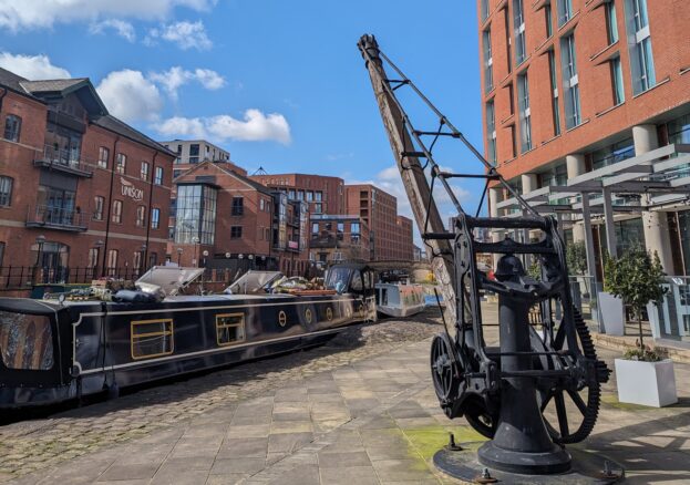 A docked narrowboat next to a heritage crane and surrounded by buildings and blue sky at Granary Wharf in Leeds