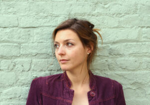 Portrait of Rebecca Watts who is white with brown hair in a purple shirt against a green wall