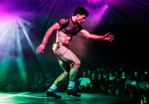 Wake at Aviva Studios: Man dances in a mesh crop top and gold shorts with purple and white irridescent lights in front of an audience.