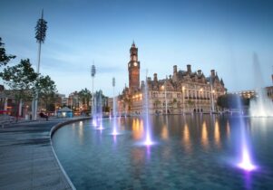 An image of a large man made pool with fountains in front of a historic building with a clock tower