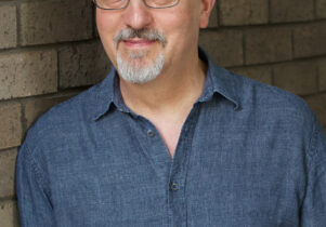 a white older man with grey hair in a blue shirt and wearing glasses leaning against a grey brick wall..