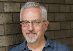 a white older man with grey hair in a blue shirt and wearing glasses leaning against a grey brick wall..