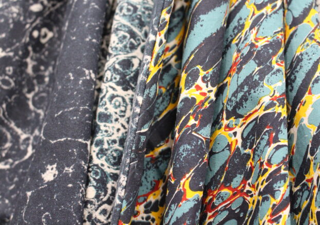 This image features a close-up of fabric with intricate, marbled patterns in various colors. The patterns resemble the swirling designs found in traditional marbling art. The fabrics display a range of tones, including dark greys, blues, teals, yellows, and red accents, creating a vibrant and textured appearance. The folds in the fabric highlight the material's flow and emphasize the rich detail of the designs.