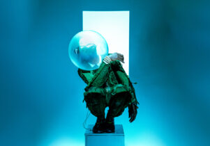 Benji Reid: Find Your Eyes at Aviva Studios: A chroreographed image a person kneeling on a pedestal in a sea green outfit, looking to the left with a bowl covering their face in front of a white light on a bluebackground