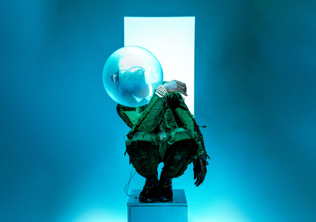 Benji Reid: Find Your Eyes at Aviva Studios: A chroreographed image a person kneeling on a pedestal in a sea green outfit, looking to the left with a bowl covering their face in front of a white light on a bluebackground