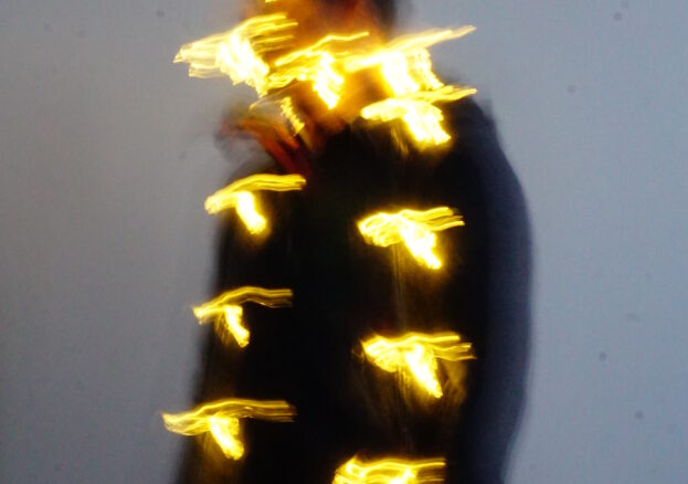 A blurred photograph of a person standing against a plain wall, wrapped in glowing string lights. The movement of the lights creates a dynamic, abstract effect, with streaks of warm yellow light appearing to float around the figure. The person's face and body are obscured due to motion blur, adding a sense of mystery and energy to the image.