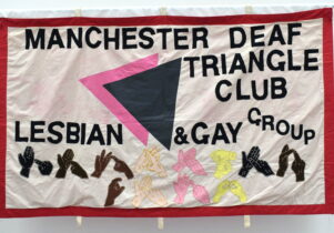 A textile banner featuring text: 'Manchester Deaf Triangle Club, Lesbian & Gay Group', and different coloured hands doing sign language. 2025 Banner Exhibition at People's History Museum